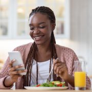 Young Smiling Black Woman Using Smartphone During Breakfast In Kitchen